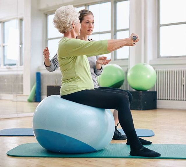 Eine Therapeuten unterweist eine Dame bei einer Übung auf einem Gymnastikball